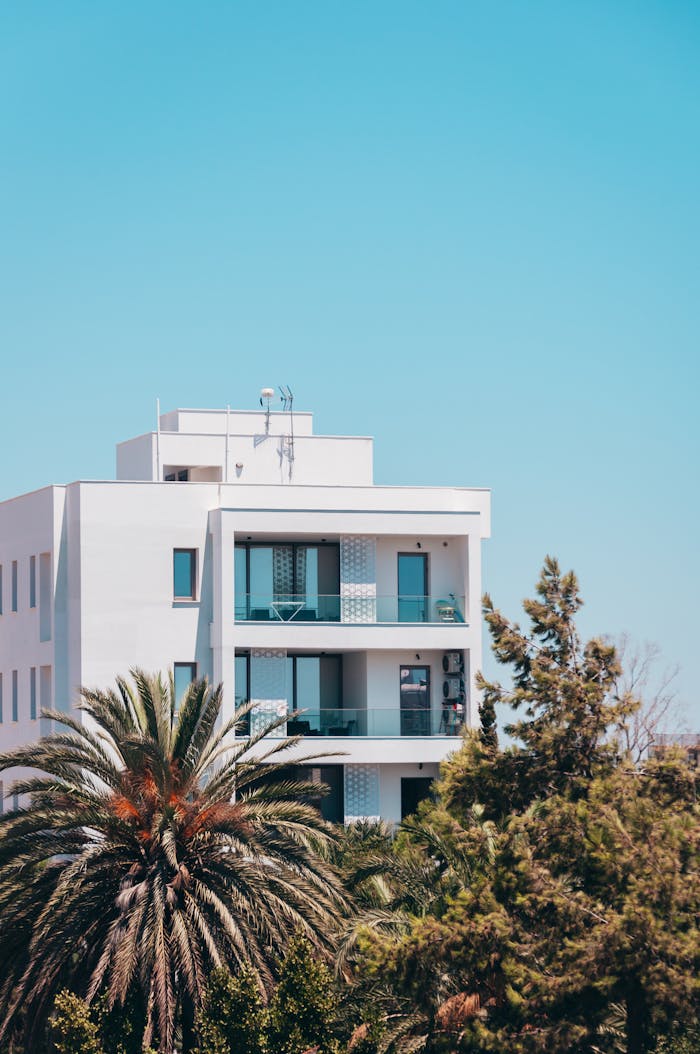 A modern white building with balconies surrounded by lush greenery and palm trees under a clear blue sky.